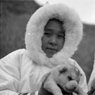 Photo de deux jeunes filles inuites vêtues de parkas blancs et tenant dans leurs mains chacune un chiot, lieu inconnu, Nunavut, juillet 1951