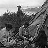 Photograph of two unidentified Aboriginal women and a young girl sitting on the ground weaving baskets in front of a tent, with a dog and a man in the background, Prince Edward Island, circa 1905