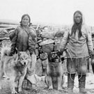 Photograph of an Inuit woman and man standing in front of stone wall with three dogs, Fullerton, Nunavut, 1904-1905